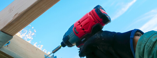 Panorama hand using cordless impact driver fasten attach hex head exterior wood screws to galvanized joist hanger support underneath rafter beam outdoor structure, fall foliage background, Texas