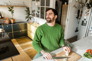 Relaxed and bearded man in green jumper looking away while holding knife and leek at home