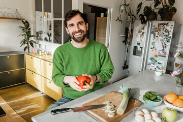 Cheerful bearded man in green jumper holding fresh bell pepper in modern kitchen at home