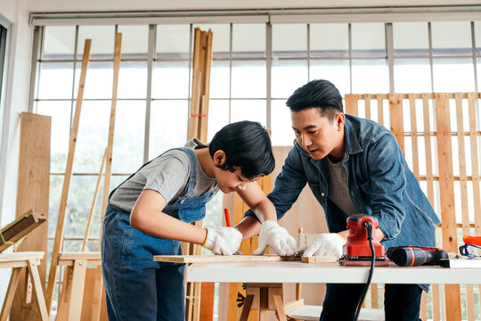 Happy Asian father and son carpentry wearing safety gloves. Father teaches his son to measure wood by measuring tape and marking with a pencil. Home workshop and Carpentry family concept.