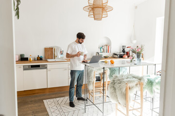 Bearded freelancer in white t-shirt and jeans holding cup of coffee and working on laptop at home