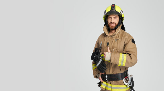 Cheerful male firefighter in uniform gesturing with thumb up. Front view of smiling bearded fireman giving like, while looking at camera, isolated on gray, copy space. Concept of work, hand gesture.