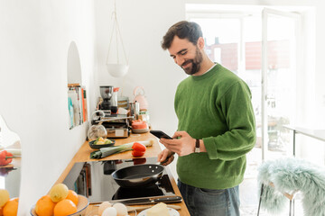 Fototapeta na wymiar Positive and bearded man in casual clothes using smartphone while cooking breakfast