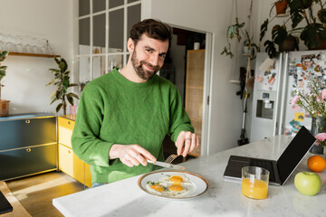 Fototapeta na wymiar Cheerful bearded man in green jumper holding cutlery near breakfast in modern kitchen at home