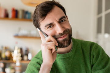 Portrait of cheerful brunette and bearded man in green jumper talking on smartphone at home