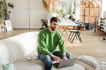 Smiling bearded tattooed freelancer in green jumper and jeans looking at camera while using laptop