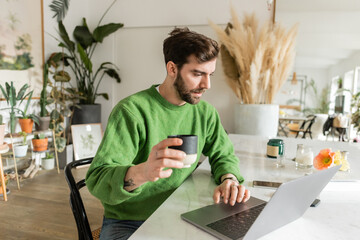 Bearded entrepreneur in green jumper holding cup of coffee and using laptop at home