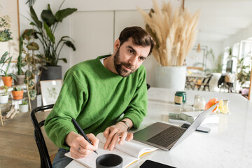 Brunette bearded freelancer in green jumper writing on notebook while working near devices on table