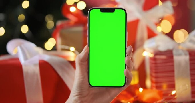 Green screen phone on Christmas decor background, close up. Chroma key screen in middle, center of static 4k footage. Smartphone with mockup on red Christmas gifts, gold garland bokeh. Copy space