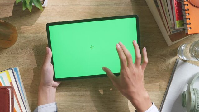 Top Down View Of Man'S Hands Using A Mock Up Green Screen Chromakey Tablet On A Wooden Office Desk Next To Books With Headphones, Pens, Keyboard, Glasses, And A Glass Of Water. Zoom In, Close Up

