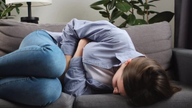 Sick young caucasian woman 25s suffering stomach pain from food poisoning lying on couch at home. Unhealthy sad girl holding belly suffering from abdomen tummy pain. Female gastritis health problem