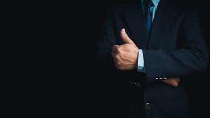 A businessman in a suit stands with his arms crossed and thumbs up on a dark background.