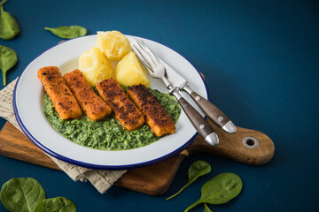 Panierte und gebratene Fischstäbchen mit Rahmspinat und Salzkartoffeln auf Emaille Teller mit Besteck, Holzbrett, Serviette, Spinat Blätter roh und dunkel blau Hintergrund