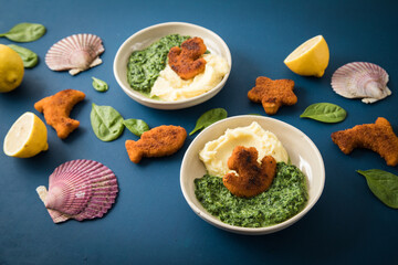 Gebratene Fischstäbchen Figuren als Fisch, Anker, Seestern und Delfin für Kinder mit Rahmspinat und Kartoffel Püree in Schale mit Zitrone, Spinat Blätter roh, Muscheln und dunkel blau Hintergrund