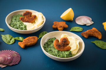 Gebratene Fischstäbchen Figuren als Fisch, Anker, Seestern und Delfin für Kinder mit Rahmspinat und Kartoffel Püree in Schale mit Zitrone, Spinat Blätter roh, Muscheln und dunkel blau Hintergrund