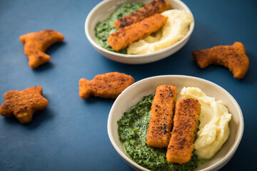Gebratene Fischstäbchen und Figuren als Fisch, Seestern und Delfin für Kinder mit Rahmspinat und Kartoffel Püree in Schale mit dunkel blau Hintergrund © Christin Klose