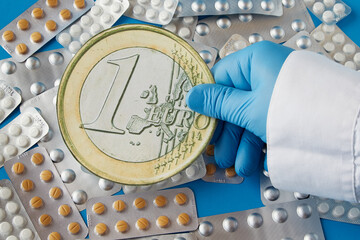 A healthcare worker's hand in a blue glove holds a large one euro coin over tablets in blisters on...