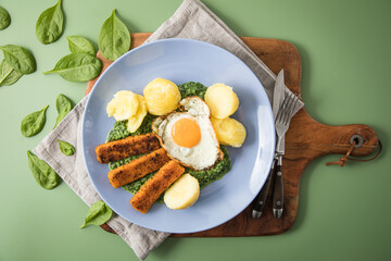 Panierte und gebratene Fischstäbchen mit Rahmspinat, Spiegelei und Salzkartoffeln auf Emaille Teller mit Besteck, Holzbrett, Serviette, Spinat Blätter roh und grün Hintergrund