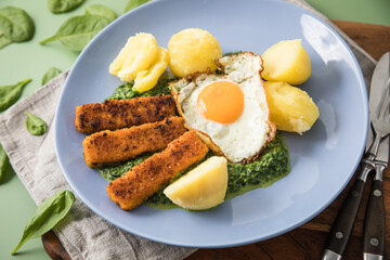 Panierte und gebratene Fischstäbchen mit Rahmspinat, Spiegelei und Salzkartoffeln auf Emaille Teller mit Besteck, Holzbrett, Serviette, Spinat Blätter roh und grün Hintergrund