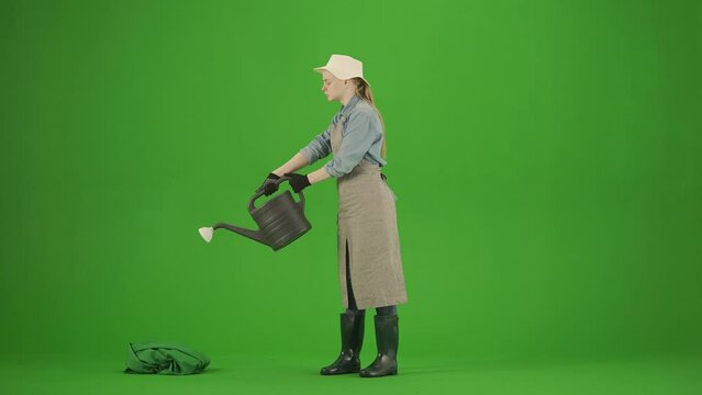 Portrait of female in apron and rubber boots on chroma key green screen. Woman gardener standing with garden watering can watering plants, smiling.