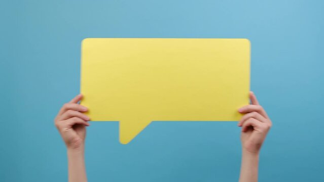 Female hands holding large empty yellow speech bubble, shaking left and right, isolated on blue color background wall in studio with copy space for promotion content or design. Dialog and idea concept