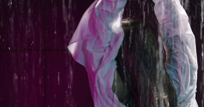 Happy young woman standing under the rain, raising hands. Wet woman dancing, water drops flow down the body. Slow motion