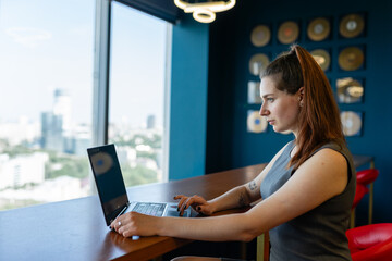 Young confident businesswoman works on laptop against panoramic window. Lady with ponytail focuses...