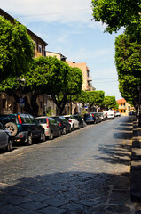 Picturesque view of narrow cobblestone street with densely parked cars. Typical Italian street with colored buildings and trees along the road. Concept of traffic and street parking in Sicily