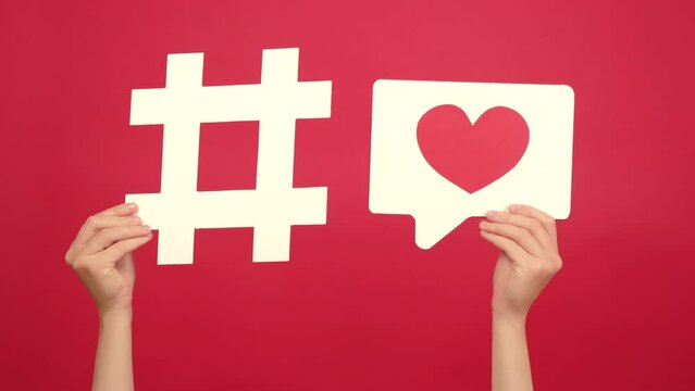 Close up of female hands holding big white hashtag sign and heart icon, isolated over red studio background wall with copy space, symbol of charity on Internet. Education, technology, online concept