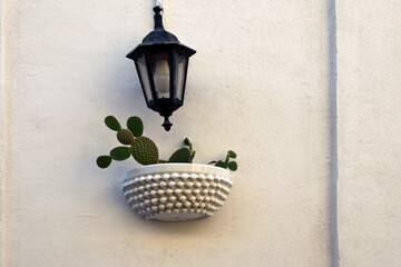 View of a light colored wall with a decorative lantern and small flower pot with a small cute cactus. Typical decoration of walls of residential building in Sicily. Santo Stefano di Camastra village