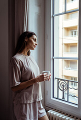 Beautiful thoughtful girl with long hair standing in bedroom looking at window drinking cofffee....