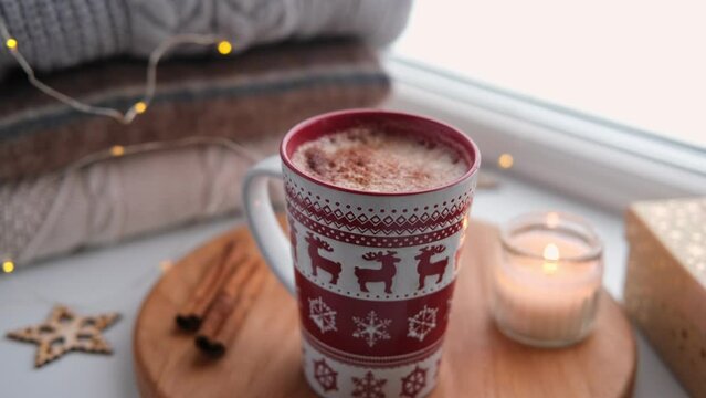 Winter windowsill still life. Red ceramic cup of hot coffee on window sill. Christmas decorations on the background. Cozy home picture. Warm woolen knitted sweaters, Burn Candle, Cookies. Stock photo	