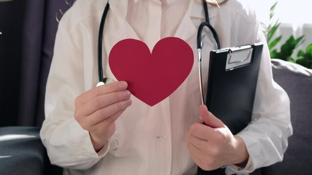 Cropped video of young woman doctor in white uniform and stethoscope holding paper red heart sitting in couch at clinic. Health insurance concept, World heart day, world health day, hypertension day