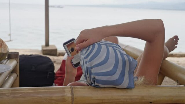 Young woman in a blue Headscarf lies on the beach, on the ocean shore, and looks at pictures on her phone. Back view. Slow Motion. 