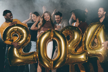 Joyful young people holding 2024 shaped gold balloons while celebrating New Year in night club
