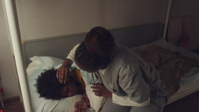Medium high angle shot of African American boy asleep in bed, mom in hospital gown coming in quietly from night shift, tucking duvet, stroking hair and kissing on cheekMedium high angle shot of Africa