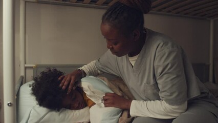 Medium shot of preteen African American boy sleeping in bedroom, mother in hospital gown walking in, tucking duvet, stroking his hair and kissing goodnight
