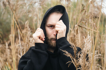 A young bearded man hides his face in a black hood, standing in the coastal reeds, the concept of...