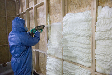 The builder in a blue suit applies a construction foam from the gun to the wall of a wooden house