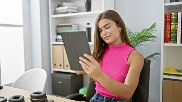 Confident young hispanic business woman grinning while working indoors; she uses her touchpad to take a beautiful office selfie