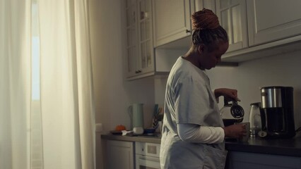 Medium shot of young African American woman in health worker uniform walking up to kitchen counter, pouring fresh coffee from coffee machine jug and drinking, while getting ready to go to work