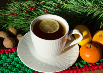 A cup of morning tea with lemon on a background of nuts, tangerines and green branches of a Christmas tree. Decorations for the New Year and Christmas.