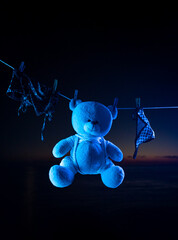 A teddy bear toy hangs on a clothesline on clothespins in neon light. tied with shibari ropes
