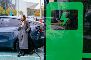 Eco friendly rechargeable EV car concept. Cheerful woman having coffee break while waiting for her...