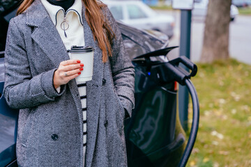 Eco friendly rechargeable EV car concept. Young woman in strip sweater standing with coffee to go,...