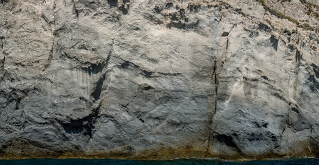Weathered seaside rock face texture with parts of green and blue water. Aged volcanic stone wall surface background pattern with cracks and scratches. Banner. Ischia Island, Italy.