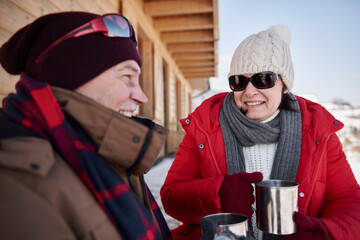 Happy mature couple with hot drinks outdoors at mountain hut in winter