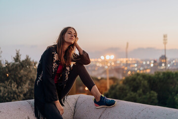 Spain, Barcelona, Montjuic, young woman sitting on a wall at dusk with city lights in background