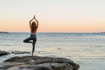 Young woman practicing yoga on the beach, doing tree pose, during sunset in calm beach, Costa...