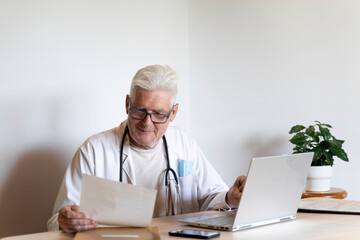 Smiling doctor looking at medical record while sitting against wall - obrazy, fototapety, plakaty